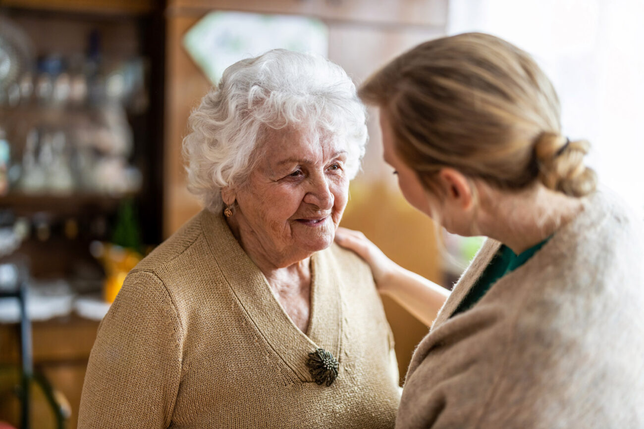 A care worker is talking to an elderly client with their hand on the client’s shoulder.